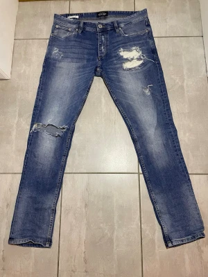 Blå slitna skinny jeans från Jack & Jones - Säljer ett par blå jeans från Jack & Jones med slitningar. Klassisk femficksmodell med knappgylf. Jeansen ha är tillverkade i bomull.
