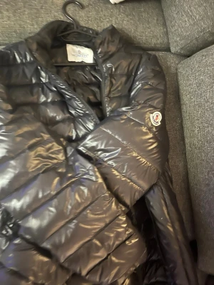 Svart dunjacka från Moncler - Snygg svart dunjacka från Moncler med glansig finish och klassisk Moncler-logga på ärmen. Jackan har quiltad design och dragkedja framtill. Perfekt för kalla dagar när du vill hålla stilen.