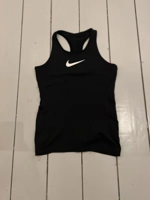 Svart träningstopp från Nike - Snygg svart träningstopp från Nike med klassisk vit logga på bröstet. Modellen är tillverkad i ett stretchigt funktionsmaterial som andas, perfekt för gymmet eller löprundan.