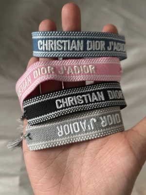 Christian Dior J'Adior tygarmband box  - Fyra snygga tygarmband från Christian Dior J'Adior i olika färger: blå, rosa, svart och grå. Armbanden har broderad text och justerbar knytning. Perfekta för att ge din outfit en exklusiv touch och visa din stil.