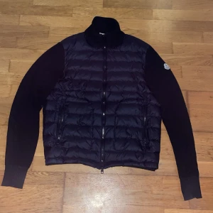 Svart cardigan jacka från Moncler - Snygg svart cardigan jacka från Moncler med quiltad front i blank polyester och stickade ärmar. Jackan har hög krage, dragkedja framtill och två fickor med dragkedja. Moncler-logga på ena ärmen ger en exklusiv touch. Pris är diskuterbart!