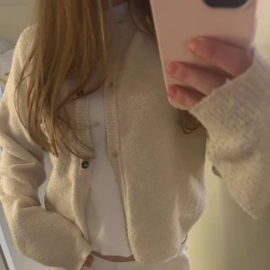 Beige stickad kofta med knappar - Mysig beige stickad kofta med långa ärmar och ribbade muddar. Koftan har en rak passform och stängs med knappar framtill. Något noprig men i överlag bra kvalité