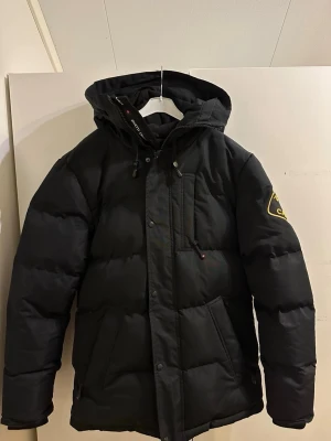 Svart pufferjacka Zavetti Canada  - Säljer en svart pufferjacka från Zavetti Canada i storlek S men passar som M. Jackan har huva, flera fickor med dragkedja och snyggt broderat märke på ärmen. Perfekt för kalla dagar med sin tjocka vaddering och stilrena look.