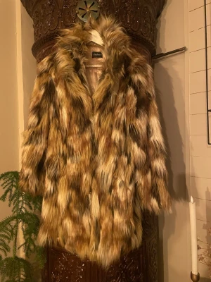 Brun pälsjacka från Miss Lola - Säljer en fluffig och trendig pälsjacka från Miss Lola i olika bruna och beige toner. Jackan har lång ärm, voluminös krage och är gjord av konstpäls i akryl och polyester. Perfekt för dig som vill sticka ut med en statementjacka i vinter.