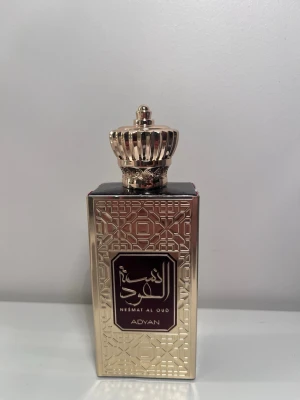 Nesmat Al Oud parfym från Adyan - Lyxig herrparfym från Adyan, Nesmat Al Oud. Flaskan är guldigt metallfärgad med orientaliskt mönster och har en dekorativ kronformad kork. Sidorna har inskriptioner och flaskan har en elegant, rektangulär form med mörka detaljer. Använd få gånger 