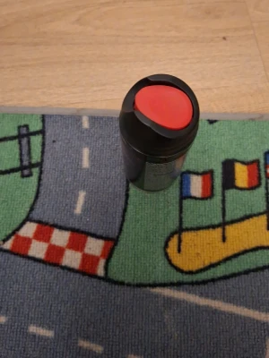 Svart termosmugg med rött lock - Praktisk svart termosmugg med ett rött lock och greppvänlig design. Muggen är tillverkad i plast och metall, perfekt för att ta med dryck på språng. Locket har en öppning för att dricka direkt utan att spilla.