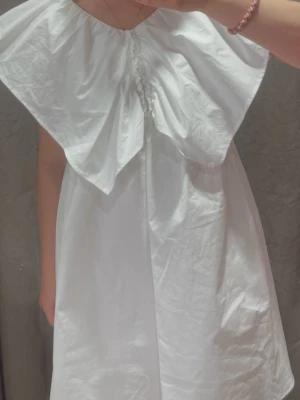Studentklänning - Säljer en vit klänning med stor volangkrage och v-ringad hals. Klänningen har en lös passform och är ärmlös, vilket ger en luftig och trendig look. Perfekt för varma dagar och enkel att styla med accessoarer. Perfekt för studenten!!