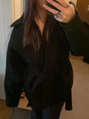 Svart kappa med bälte och fickor - Stilren svart kappa med bälte i midjan och stora fickor framtill. Klassisk krage och långärmad modell som ger en snygg och avslappnad look. Perfekt för höst och vinter, lätt att matcha till olika outfits.