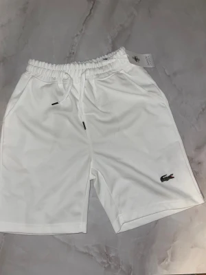 Vita shorts från Lacoste med snörning - Snygga vita shorts från Lacoste med klassisk krokodillogo broderad på benet. Shortsen har elastisk midja med snörning och två sidofickor. Perfekta för en clean och sportig look.