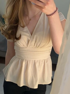 Beige omlottopp - Snygg beige topp med omlott-skärning och bred midjedetalj. Toppen har korta ärmar och volang nedtill som ger en feminin siluett. Perfekt att styla med jeans eller kjol. Toppen är i fint skick utan några skavanker.