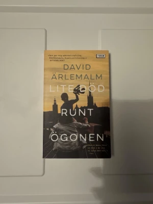 Lite död runt ögonen - En gripande roman av David Ärlemalm som utspelar sig i Stockholm. Omslaget har en siluett av en staty mot en gul och svart bakgrund med stadssilhuett. Boken handlar om relationer, förlust och kampen för att hitta tillbaka till livet.