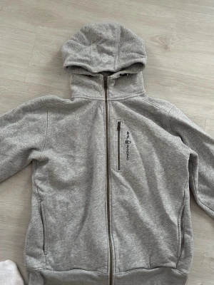 Grå hoodie från Sail Racing - Mjuk och skön grå hoodie från Sail Racing med dragkedja framtill och broderad logga på bröstet. Hoodien har en bröstficka med dragkedja, två sidofickor och vit fleece på insidan för extra komfort. Perfekt för chill dagar eller när du vill ha en sportig look.