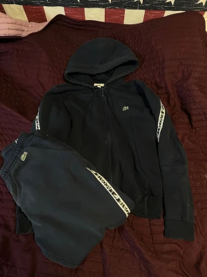 Svarta mjukisbyxor från Lacoste - Säljer ett par svarta mjukisbyxor från Lacoste med klassisk logga på benet och vita sidoremsor med Lacoste-tryck. Byxorna har resår i midjan och är perfekta för chill dagar. Matchar snyggt med hoodie eller t-shirt. Koftan är S och byxorna är M