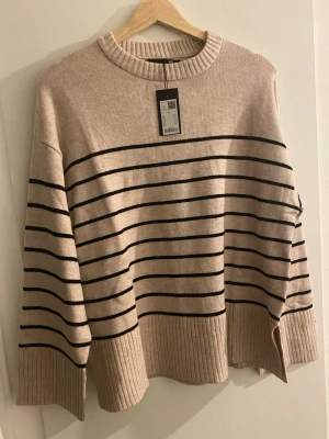 Beige stickad tröja med svarta ränder - Säljer en beige stickad trendig tröja med tunna svarta horisontella ränder. Tröjan har rund halsringning, breda ribbade muddar och en avslappnad passform. Perfekt för dig som gillar klassisk och enkel stil. Prislapp kvar ✨