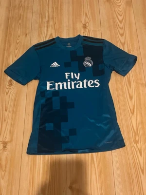 Blå Real Madrid matchtröja Adidas - Säljer en blå Real Madrid matchtröja från Adidas med Fly Emirates-tryck på bröstet. Tröjan har korta ärmar, svart pixelmönster på framsidan och klubbmärke på vänster bröst. Tillverkad i lätt funktionsmaterial, perfekt för fotbollsträning.