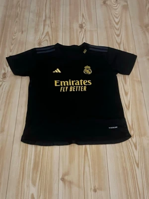 Real Madrid Bellingham svart tröja - Svart Real Madrid-tröja från Adidas med guldiga detaljer, klubbmärke och sponsortryck framtill. Nummer 5 och Bellingham på ryggen. Korta ärmar och rund hals. Perfekt för fotbollsfans som vill sticka ut på planen eller läktaren.