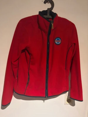 Röd fleecejacka från Mountain Horse - Röd fleecejacka från Mountain Horse med hel dragkedja och ståkrage. Jackan har två fickor med dragkedja och en broderad logga på bröstet. 