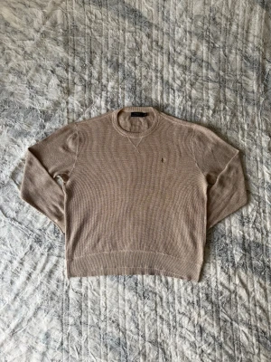 Beige stickad tröja från Polo Ralph Lauren - Beige stickad tröja från Polo Ralph Lauren med rund halsringning och diskret broderad logga på bröstet. Storlek L. Tröjan har ribbstickade muddar och är tillverkad i ett mjukt, grovstickat material. Perfekt för lager-på-lager och en avslappnad stil. Tröjan är i nyskick och är knappt använt. Nypris är 2399kr.