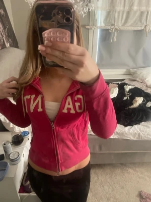 Rosa zip hoodie från GANT - Säljer en rosa hoodie från GANT med dragkedja framtill och stora vita bokstäver på bröstet. Hoodien har fickor på sidorna och en klassisk huva. Perfekt för en chill och sportig look. Materialet känns mjukt och bekvämt. har små fläckar dock så är de inget man lägger märke till