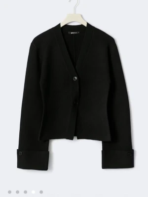 Svart cardigan med knappar från Gina - Stilren svart cardigan med två knappar framtill och breda uppvikta muddar med knappdetalj. Djup V-ringning och rak passform ger en modern look. Perfekt att slänga över en t-shirt för en enkel men snygg outfit. Den är i ett bra skick, bara att hålet till knappen längst upp kan ha förstorats lite. Men det är inget man tänker på eller nåt som förstör tröjan. Skriv om mer bilder eller något sånt 