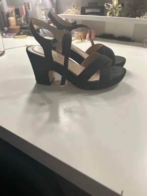 Svarta sandaler med klack från Savita - Snygga svarta sandaler från Savita med breda remmar och justerbart spänne runt ankeln. Skorna har öppen tå och hög klack, tillverkade i mocka-liknande material. Perfekta för dig som vill ha både stil och komfort.