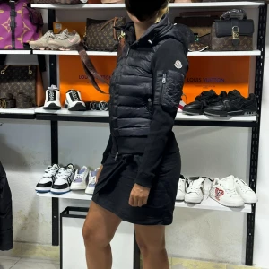 Svart dunjacka från Moncler XS/S - Snygg svart dunjacka väst från Moncler med quiltad front, huva och dragkedja. Jackan har långärmade stickade ärmar och Moncler-logga på ärmen. Perfekt för kyliga dagar och ger en sportig men stilren look.