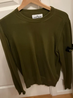 Olivgrön stickad tröja Stone Island - Snygg olivgrön stickad tröja från Stone Island med rund halsringning och klassisk logotyp-patch på vänster ärm. Tröjan har långa ärmar och ribbade muddar vid ärmslut och nederkant. Perfekt för dig som gillar streetwear och stilren design.