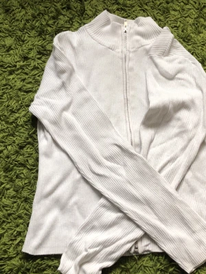 Vit ribbad ziptröja med hög krage - Snygg vit ribbad tröja med dragkedja framtill och hög krage. Långärmad modell i stretchigt material som sitter tajt och ger en clean look. Perfekt att matcha med jeans eller kjol för en enkel men stilren outfit.