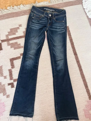 Mörkblå bootcut jeans American Eagle - Snygga Lågmidjade mörkblå bootcut jeans från American Eagle med klassisk femficksdesign och fransade benslut. Jeansen har en lätt tvättad look och är tillverkade i mjukt denimtyg med stretch för extra komfort.