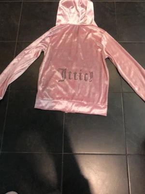 Rosa velour hoodie från Juicy Couture - Säljer en ikonisk rosa velour hoodie från Juicy Couture med dragkedja framtill och fickor. På ryggen står det 'Juicy' i glittrig text. Hoodien har huva och långa ärmar, perfekt för en avslappnad och trendig look.