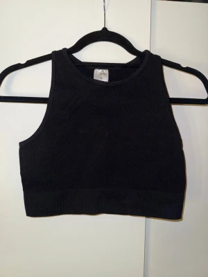 Svart ribbad croppad linnetopp - Snygg svart croppad linnetopp med ribbad kant. Toppen är ärmlös och har en hög halsringning, perfekt att styla till jeans eller kjol. Enkel och stilren design som passar till många outfits.