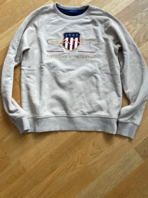 Beige sweatshirt från Gant - Snygg beige sweatshirt från Gant med broderad logga och texten 'American Sportswear' framtill. Tröjan har rund halsringning, ribbade muddar och klassisk collegekänsla. Perfekt för en avslappnad och sportig stil.