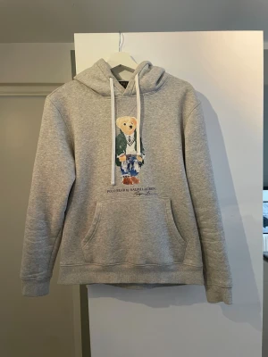 Grå hoodie Polo Bear Ralph Lauren - Säljer en grå hoodie från Polo Ralph Lauren med den ikoniska Polo Bear-trycket framtill. Hoodien har huva med vita snören och en stor magficka. Mjuk bomullsblandning och avslappnad passform, perfekt för en chill och stilren look.