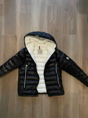 Svart dunjacka med huva från Moncler - Snygg svart dunjacka från Moncler med vit insida och huva. Jackan har två stora dragkedjefickor framtill, quiltad design och Moncler-logga på ärmen. Perfekt för kalla dagar och ger en modern, sportig look.