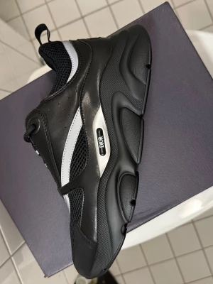 Dior bb22 pris kan diskuteras  - Chunky svarta Dior sneakers med silvriga detaljer och meshpaneler. Skorna har en kraftig sula, snörning och tydlig Dior-logga på sidan. Kombination av skinn och mesh ger en sportig och modern vibe. Perfekta för dig som vill sticka ut med exklusiva sneakers.
