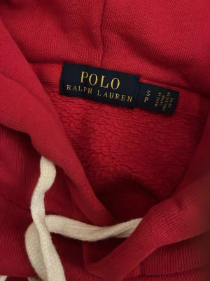 Röd hoodie från Polo Ralph Lauren - Snygg röd hoodie från Polo Ralph Lauren med klassisk broderad polospelare på bröstet. Hoodien har vit dragsko och mjukt material på insidan. Perfekt för en avslappnad och trendig stil.