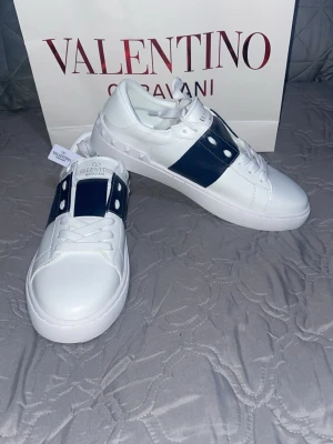 Valentino Garavani vita sneakers - Snygga vita sneakers från Valentino Garavani med en mörkblå detalj på sidan och klassisk snörning. Skorna har en platt sula och är tillverkade i skinn med diskreta nitar längs kanten. Perfekta för dig som gillar stilrena och exklusiva sneakers. Helt nya