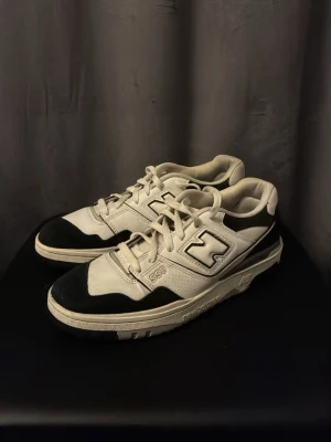 New Balance 550 svartvita sneakers - Snygga New Balance 550 sneakers i svart och vitt med klassisk retrodesign. Skorna har snörning, rund tå och platt sula. Perfekta för dig som gillar streetwear och vill ha en stilren look med tydlig logga på sidan.