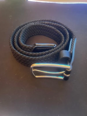 Svart flätat bälte med silverfärgat spänne - Snyggt svart flätat bälte med ett stilrent silverfärgat metallspänne. Bältet har detaljer i svart läder vid spännet och passar perfekt till jeans eller chinos. Enkel och modern design som funkar till många olika outfits.