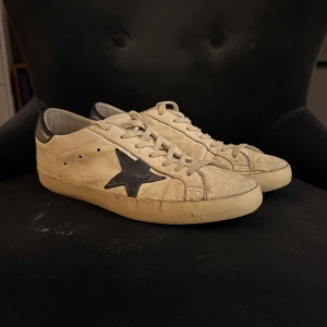 Golden Goose skor - Golden Goose Superstar sneakers i ljusbeige skinn med mörkblå stjärna på sidan. Klassiska superstarts. Snyggt slitna detaljer och retrovibe, perfekt för dig som gillar streetstyle. Nypris 5500kr. Slitningar i hälen men något som inte syns allt för mycket.