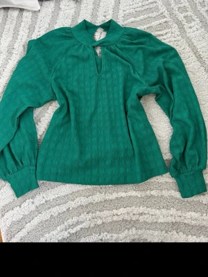 Grön blus med struktur och nyckelhålsringning - Snygg grön blus med långärmade puffärmar och breda muddar. Blusen har en unik nyckelhålsringning framtill och är tillverkad i ett strukturerat tyg som ger en cool känsla. Perfekt för dig som vill sticka ut med färg och form.