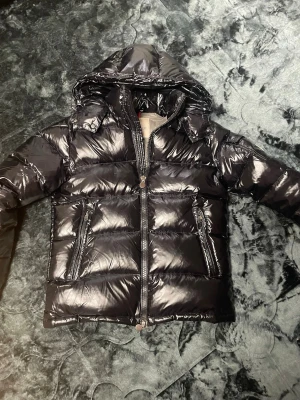 Svart glansig dunjacka från Moncler - Snygg svart glansig dunjacka från Moncler med huva och dragkedja framtill. Jackan har två framfickor med dragkedja och quiltad design. Insidan är ljusgrå och har en innerficka med dragkedja samt Moncler-logga i nacken.