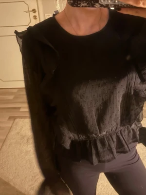 Svart blus med volangdetaljer - Snygg svart blus med transparenta långärmar och volang vid midjan. Blusen har diskret struktur i tyget och feminina detaljer vid axlarna. Perfekt för dig som gillar en trendig och stilren look.💕