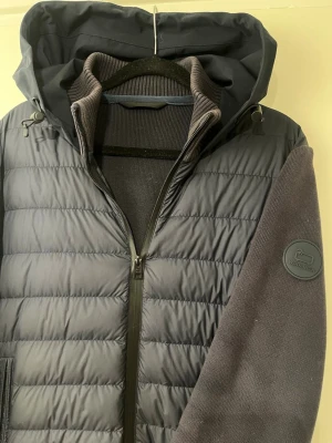 Woolrich cardigan  - Snygg mörkblå cardigan från Woolrich med quiltad front och huva. Jackan har stickade ärmar och muddar, dragkedja framtill och diskret logotyp på ärmen. Perfekt för kyliga dagar när du vill ha både stil och komfort. Det är storlek S men passar även xs, normal Woolrich storlek!