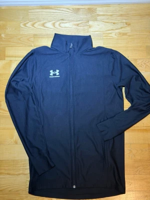 Svart träningströja från Under Armour - Snygg svart träningsjacka från Under Armour med hel dragkedja och hög krage. Jackan har logga på bröstet och är tillverkad i ett lätt och stretchigt material som passar perfekt till träning och sportaktiviteter.