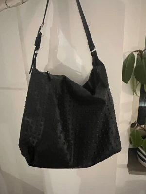 Svart axelväska med struktur - Cool svart axelväska med justerbart axelband och unik prickig struktur på hela väskan. Väskan är rymlig och har en mjuk, slouchy form som gör den perfekt för att bära med sig allt man behöver. Passar dig som gillar en stilren men ändå lite edgy look.