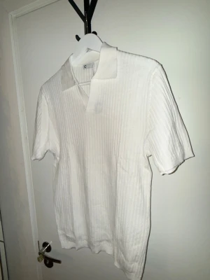 Vit ribbad polotopp med krage 100% bomull - Vit ribbad polotopp med kort ärm och klassisk krage. Toppen har en stilren och enkel design med diskreta vertikala ränder i materialet. Perfekt för en clean och fräsch look.