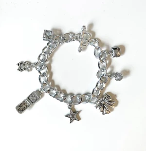 Silvrigt charmarmband med berlocker - Säljer ett chunky armband i silverfärgad metall med flera berlocker. Länkarna är grova och armbandet stängs med en T-lås. Perfekt accessoar för dig som gillar statement-smycken🤗du får med en liten tygpåse.
