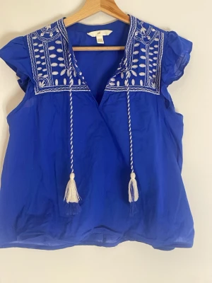 Blå broderad blus från H&M - Blå blus från H&M med vita broderier på axlarna. Har använt ett fåtal gånger så mycket bra skick. Säljer för att den inte blir till användning.