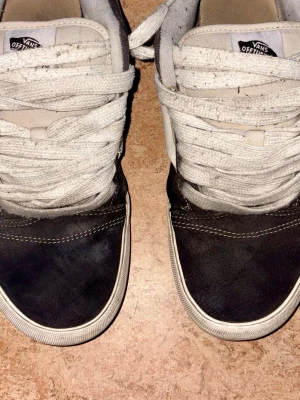 Svarta Vans sneakers med vita snören - Klassiska Vans sneakers i svart mocka och textil med vita snören och vit sula. Skorna har rund tå, platt sula och tydliga sömmar för en clean streetstil. Perfekta för dig som gillar enkel och tidlös design.